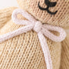 Close-up of the hand-knit Cuddle + Kind Baby Honey Bear with a lavender bow. The heirloom-quality beige teddy bear features a partially visible face with a stitched black nose and mouth.