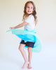 A young child with long, curly hair twirls joyfully, barefoot on a light floor, wearing a white t-shirt, dark shorts, and Sarah's Silks Enchanted Playsilk - Sea as a flowing blue accessory against a neutral background.