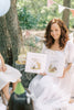 A woman in a white dress reads Winnie-the-Pooh: Finding Feelings to kids outside, surrounded by trees, balloons, pastel bunting, and a candle-topped cake on a stump.