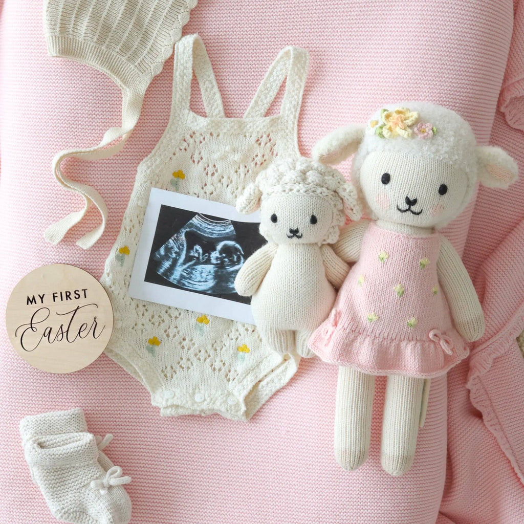 A flat lay of baby items on a pink blanket, featuring a knitted romper, bonnet, booties, an ultrasound photo, a wooden "My First Easter" sign, and Cuddle + Kind Eleanor the Lamb hand-knit doll.