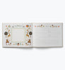 The Storybook Embroidered Baby Book features an open layout with an illustrated animal and nature border on the left page, while the right page offers lined sections for writing memories and notes, all set against a crisp white background.