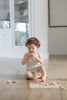 A toddler dressed in light colors sits on a wooden floor, playing with the Farm Animals Wooden Puzzle: A Tabitha Paige Puzzle and holding a puzzle piece up to their face.