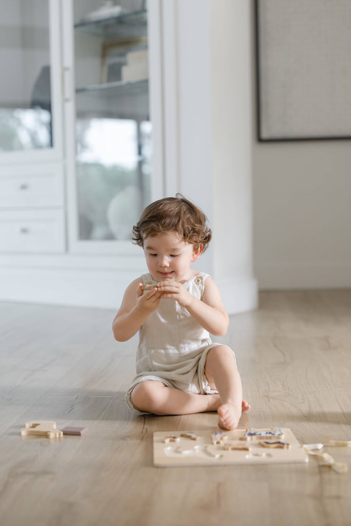 A toddler dressed in light colors sits on a wooden floor, playing with the Farm Animals Wooden Puzzle: A Tabitha Paige Puzzle and holding a puzzle piece up to their face.