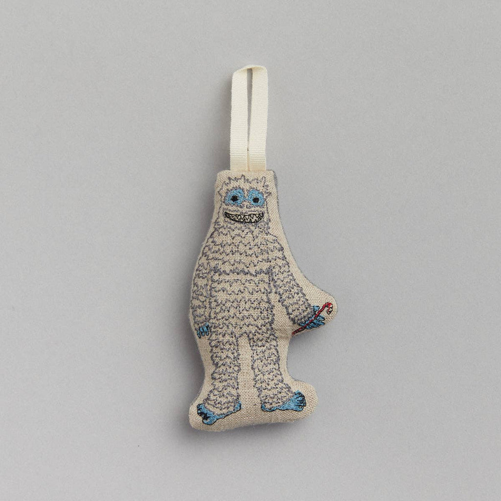 Ornament Description: The Yeti with Candy Cane Ornament features a plush design in the shape of a yeti, complete with white fur, blue facial details, and a joyful expression. It holds a small red candy cane in one hand and includes a white loop on top for easy hanging. The ornament is set against a plain gray background.