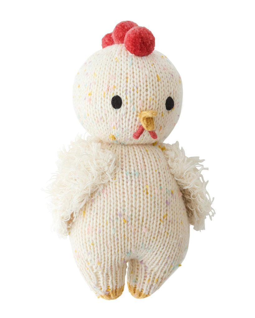 The Cuddle + Kind Baby Chicken is a hand-knit, heirloom-quality doll crafted in Peru, featuring white yarn, fuzzy wings, a yellow beak, red comb, small black eyes, and multicolored specks on its body.