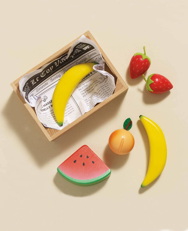 The Smoothie Fruit Wooden Market Crate (ships in one week) features wooden bananas, strawberries, a peach, and a watermelon slice on a beige surface—ideal for imaginative play or early learning. Some pieces sit outside the decorative wooden crate.