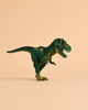 The Schleich Tyrannosaurus Rex Dinosaur Toy with Movable Jaw, made from green plastic, stands on a beige surface with a plain beige background.