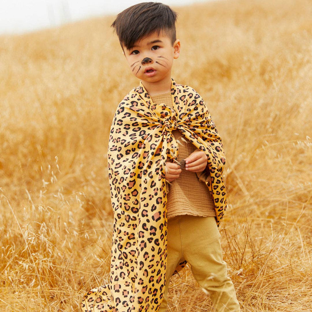 A young child stands in a dry field wearing Sarah's Silks Animal Print Playsilk - Cheetah as a cape and cat-inspired face paint, dressed in tan clothing and looking slightly to the side.
