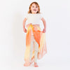 A young child stands smiling, wearing a white shirt and leggings with Sarah's Silk Enchanted Playsilk in Desert colors tied around the waist, creating a flowing cape. This simple addition sparks imaginative play against the plain white background.