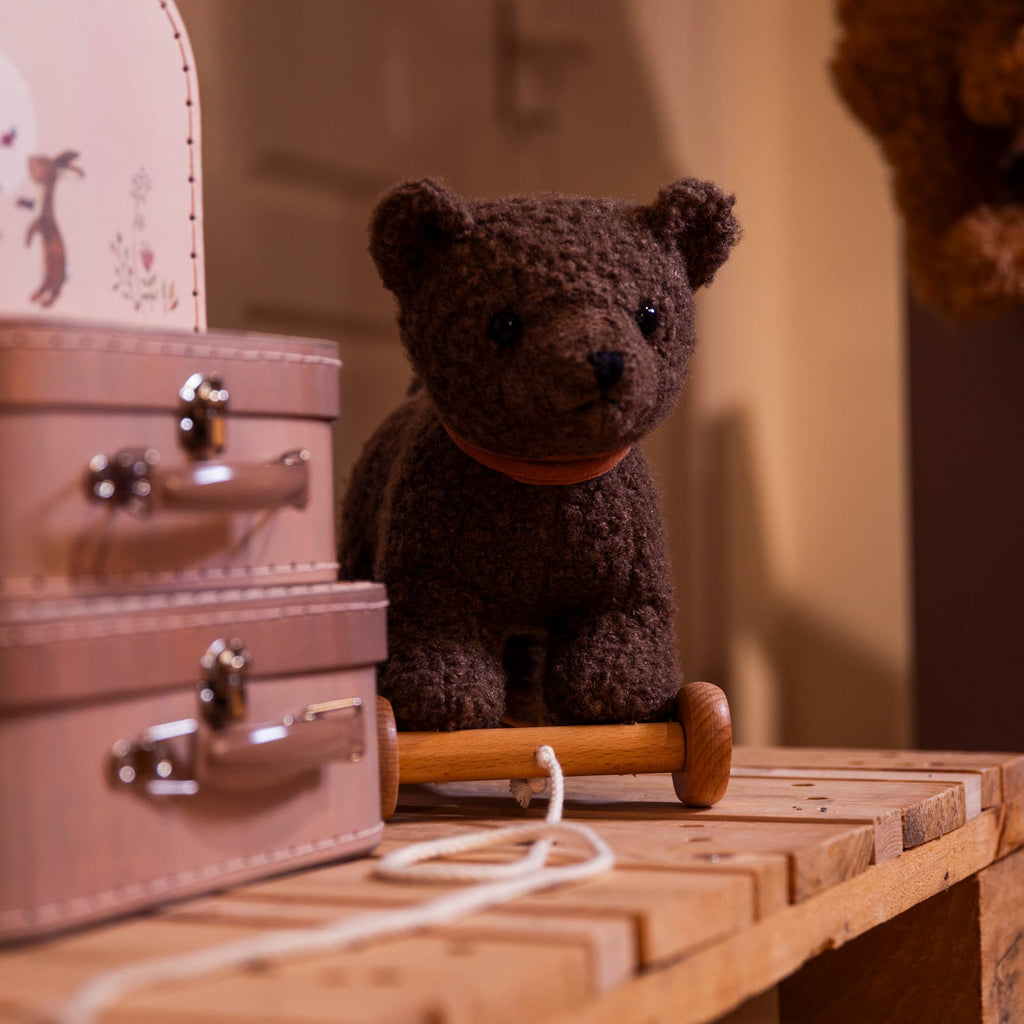 Pull-Along Raoul, a plush brown bear toy by Egmont Toys, sits on wooden wheels with a pull string atop a wooden table next to two small, stacked suitcases with metal clasps. The softly lit background is gently out of focus.