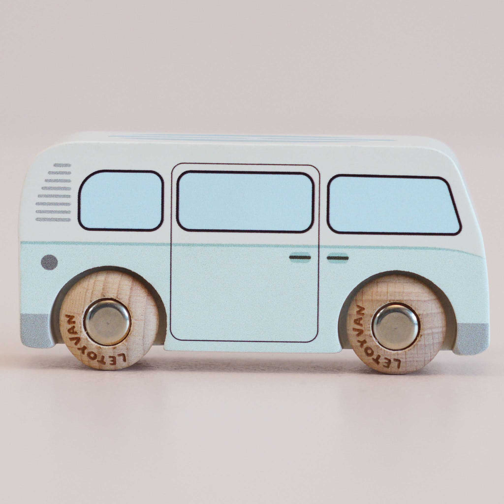 A pastel blue wooden toy van labeled “No. 29 GT160 Daisy | Made in England,” with simple black outlines and wooden wheels, evokes a charming retro design and hints at slow adventures, all while sitting on a neutral background.