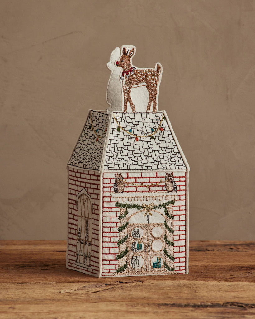 The Coral & Tusk Home for the Holidays Tissue Box Cover is a decorative, embroidered house featuring a deer on the roof, brick walls, festive garlands, owls, and a door—shown on a wooden surface with a plain background.