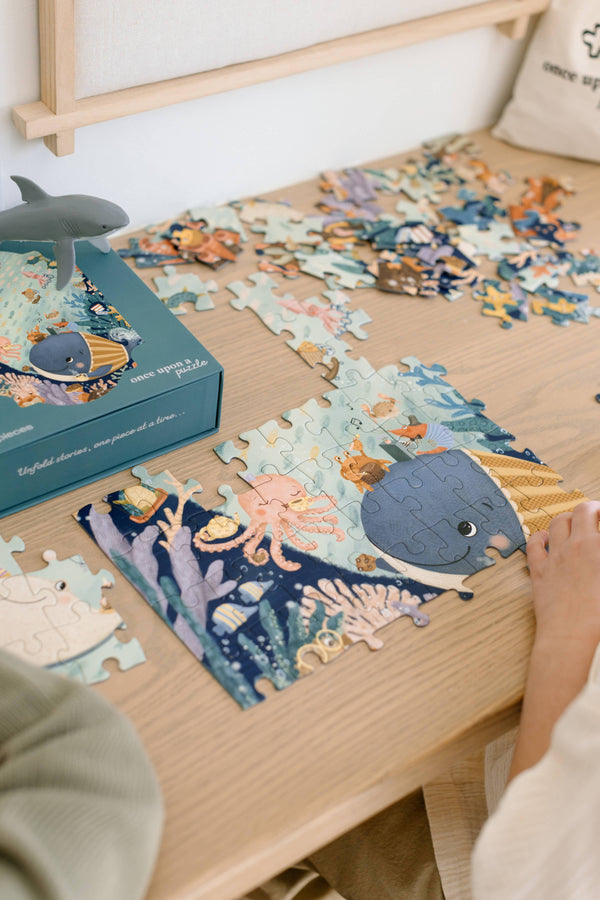 A person assembles the 100-piece "Tales from the Deep" puzzle, designed for ages 5+, on a wooden table. The ocean-themed puzzle features sea animals like a blue whale and fish, with its illustrated box and a toy shark nearby.