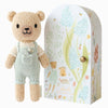 The Cuddle + Kind Tiny Charlie the Honey Bear, a hand-knit doll in light green overalls, stands next to a decorative box with pastel bears, flowers, bees, and “charlie the honey bear.” Lovingly handmade in Peru.