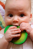 A baby with big brown eyes chews on the Arnold the Avocado Teething Toy, made of natural rubber, while wearing a white knitted outfit.