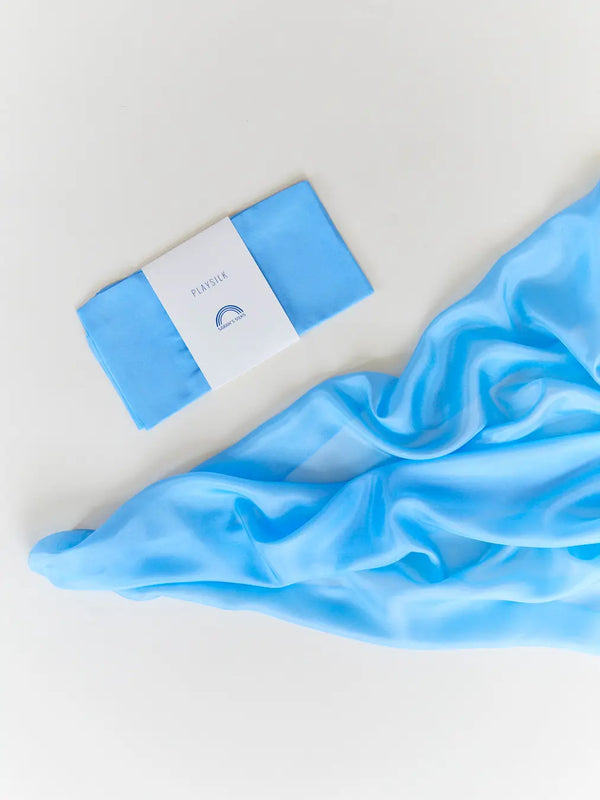 A Sarah's Silk Playsilk - Sky Blue lies partially unfolded next to its packaging on a clean, white surface. The packaging features a simple design with the word "classic" and a crescent moon logo, suggesting its