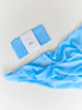 A Sarah's Silk Playsilk - Sky Blue lies partially unfolded next to its packaging on a clean, white surface. The packaging features a simple design with the word "classic" and a crescent moon logo, suggesting its