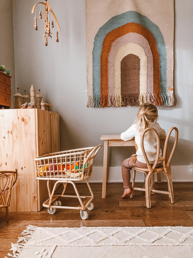 A young child sits at a small wooden table in a cozy room, facing a colorful woven rainbow tapestry on the wall. A miniature shopping cart filled with Toddler Rattan Bunny Chairs is beside them.