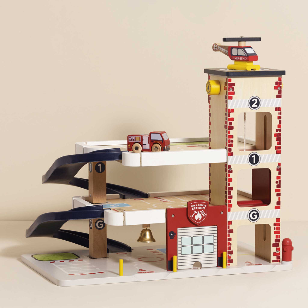 The Fire & Rescue Garage is a wooden toy set with ramps, a fire engine, chief’s car, helipad with red helicopter, numbered multi-level floors, and a base featuring road markings.