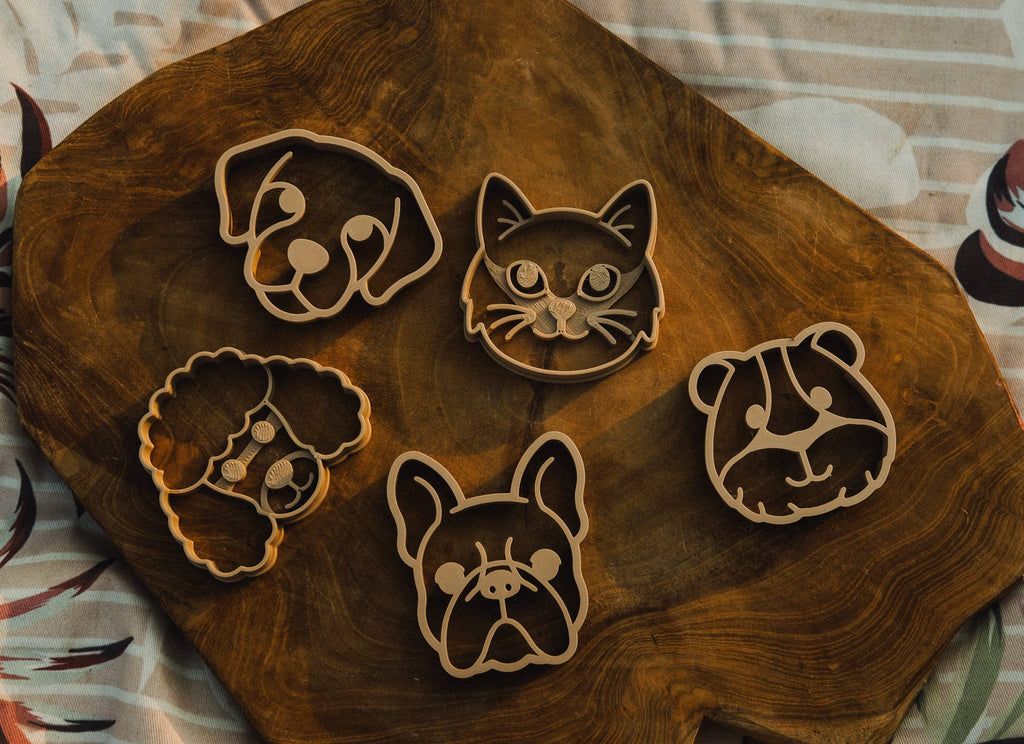 Five animal-shaped cutters—dog, cat, poodle, bulldog, and bear—are arranged on a wooden board atop a patterned cloth background, making the Pet Friends Faces Cutter Set - Eco Playdough Tool perfect for inspiring creativity and sensory play.