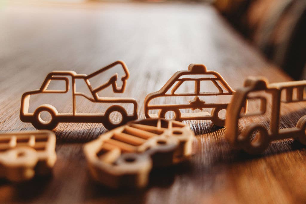 The Service Vehicle Cutter Set - Eco Playdough Tool is displayed on a wooden surface in warm natural light alongside wooden toy vehicles—a tow truck, police car, and fire truck—perfect for inspiring sensory play or pairing with transport-themed accessories.