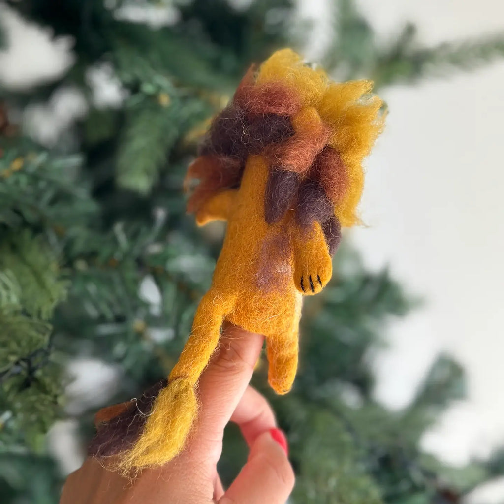 The Fluffy Lion Finger Puppet, handmade in Nepal from felted wool, displays a yellow body complemented by a multicolored mane in shades of brown and orange. It perches on an outstretched finger against a backdrop of blurred green foliage, evoking a natural setting. Ideal as a distinctive storytelling prop.