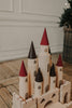 Discover the enchantment of the Great Kingdom Building Set, a wooden toy castle with towering conical spires in vibrant red and earthy brown. Meticulously crafted with smooth edges and painted using non-toxic materials, this captivating construction is beautifully showcased on a wooden floor, set against a white wall and complemented by a basket of greenery for an idyllic setting.