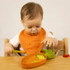 A baby wearing an orange bib sits at a table, playing with food using a spoon and their non-toxic dinnerware. There is food smeared on the baby’s face and hands, with the Cathy the Carrot Plate & Spoon Set placed in front of them.