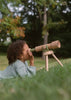 A young child with curly hair lies on the grass, looking through the Wooden Tripod Telescope – 15x Zoom, surrounded by trees and greenery.