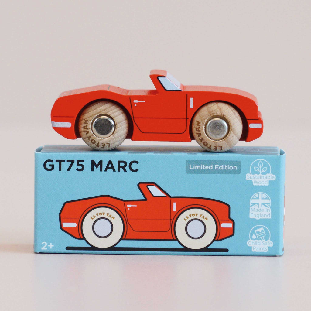 A racing red wooden toy car rests on a blue box displaying an illustration of the classic design, the product name “No. 13 GT75 Marc | Made in England,” and icons denoting sustainable wood, made in England, and child-safe paint.