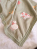 The Organic Cotton Knit Baby Blanket - Farm Animals is a soft green blanket with embroidered animals—a pink and white cow, a sheep, and part of a yellow animal—shown on a white furry surface.
