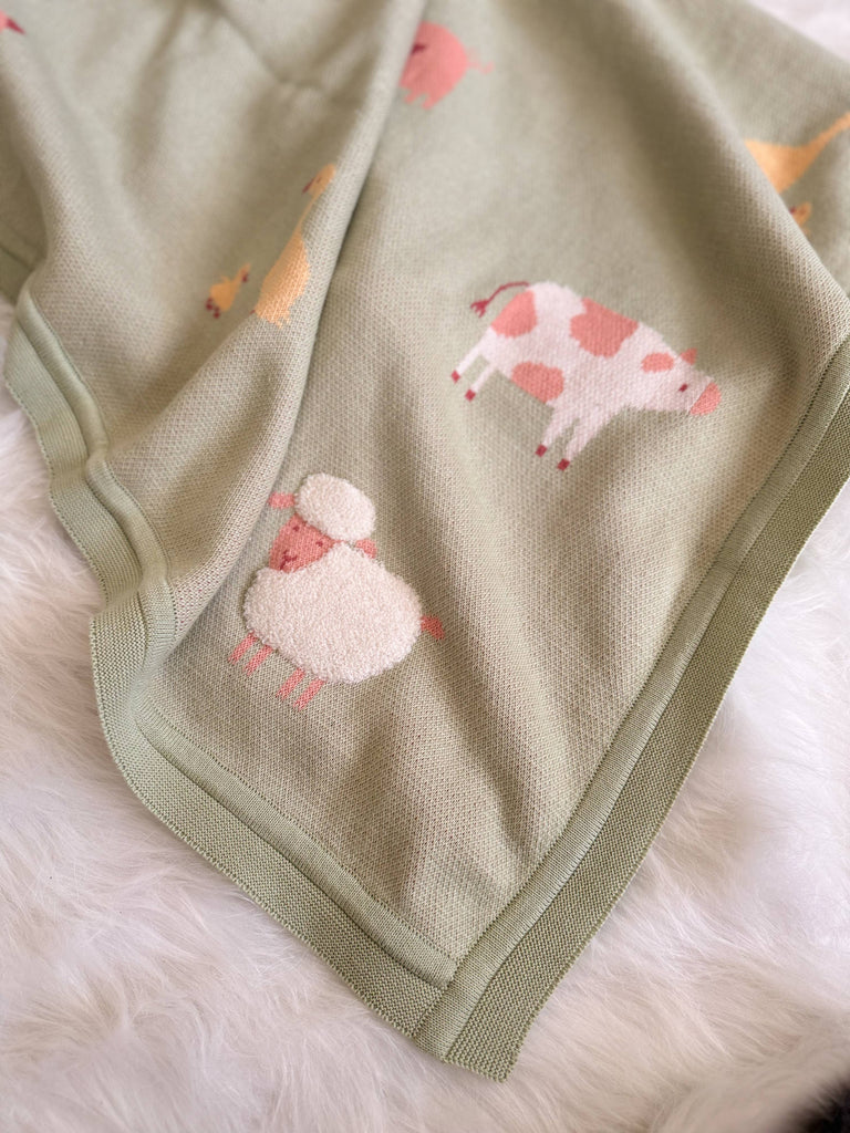 The Organic Cotton Knit Baby Blanket - Farm Animals is a soft green blanket with embroidered animals—a pink and white cow, a sheep, and part of a yellow animal—shown on a white furry surface.