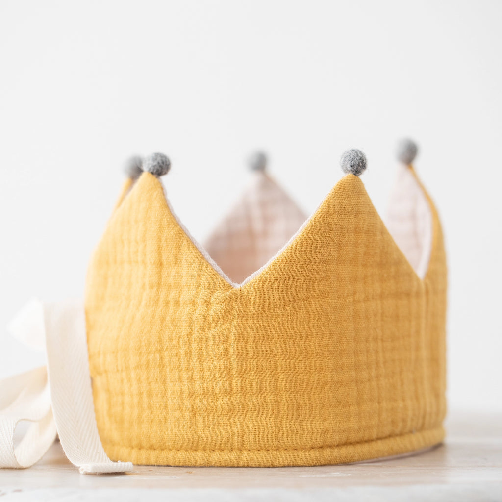 A soft, yellow fabric Reversible Crown handmade in Spain with pointed peaks topped with gray pom-poms, set against a light, neutral background. The crown features an adjustable tie at the back.