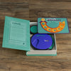 A Board Game: Catch Phrase Vintage Bookshelf Edition is open on a wooden floor, showing its light green box with the game board, instruction sheet, and circular electronic game unit inside.