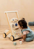 A toddler in blue overalls kneels on a wooden floor, playing with the Wooden Activity Walker, which features various interactive shapes and sensory toys. Colorful blocks are scattered nearby.