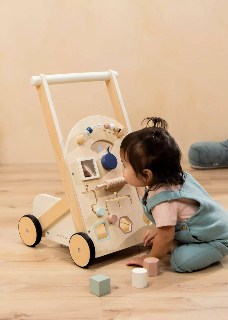 A toddler in blue overalls kneels on a wooden floor, playing with the Wooden Activity Walker, which features various interactive shapes and sensory toys. Colorful blocks are scattered nearby.