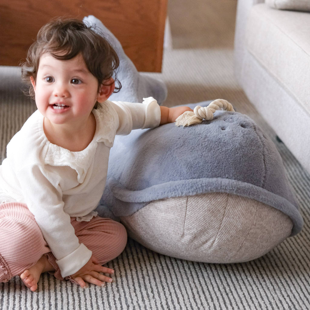 A smiling toddler with curly hair sits on the floor, wearing a white top and pink pants, resting one hand on the large Ride-On Whale Pouf, which doubles as a cozy floor cushion.