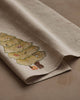 The Coral & Tusk Peek a Tree Tea Towel is a beige linen cloth embroidered with a woodland Christmas tree adorned with ornaments and gifts, capturing the magic of Christmas morning on a neutral backdrop.