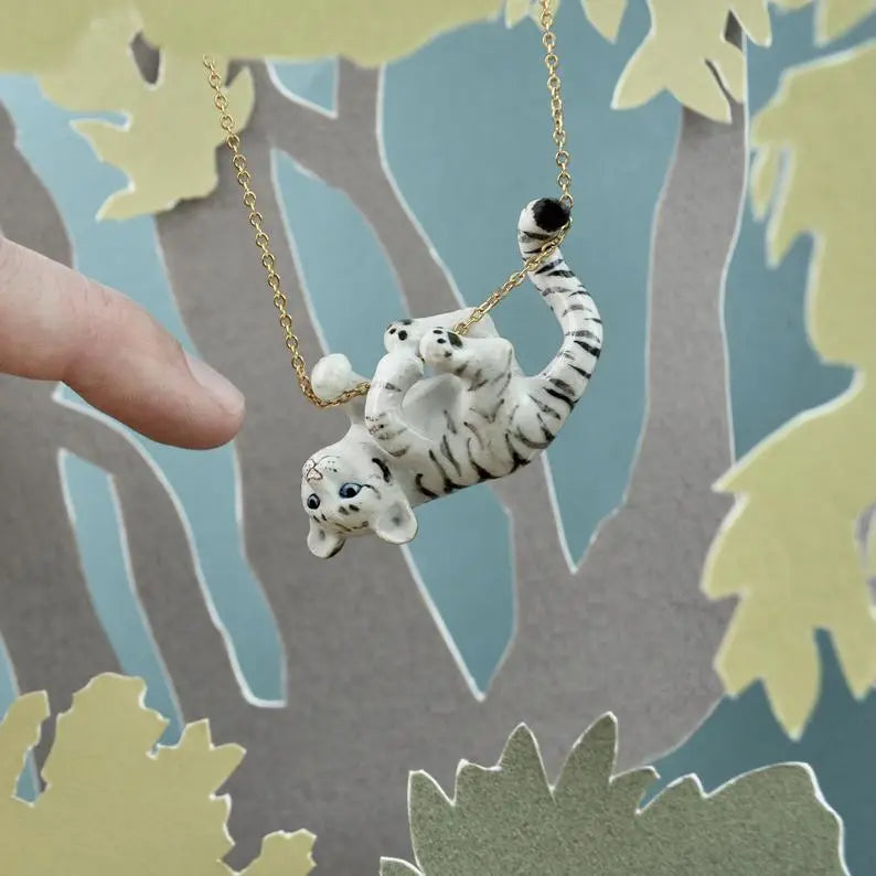 A hand holds a White Tiger Cub Necklace featuring a playful, hand-painted ceramic white tiger, suspended on a 24k gold plated chain, swinging from a black and white striped branch against a leafy backdrop.
