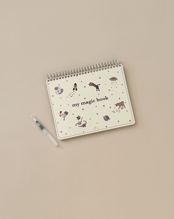 A beige spiral-bound "My Magic Water Book - Sleet" lies on a matching beige surface. The cover features whimsical illustrations of various animals and objects. A white and gray refillable water brush pen is placed to the left of the book.