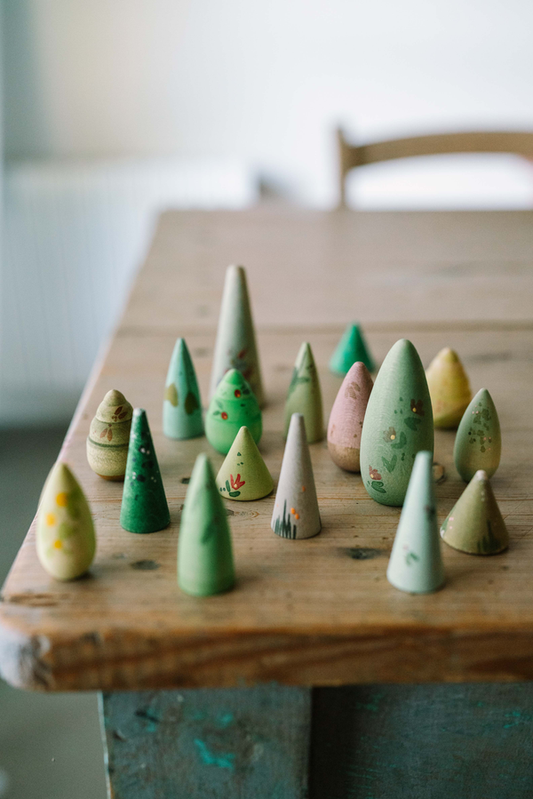 The Grapat Play Set - Forest Tree features small, cone-shaped sustainable wooden trees painted with non-toxic green dyes and patterns, arranged on a rustic wooden table with a blurred chair and background.