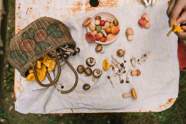 The Grapat Play Set - Mushroom Grove features a woven basket of assorted mushrooms spilling onto a white cloth on a rustic table, with both real and hand-painted wooden mushrooms—ideal for open-ended play—and a hand picking up a yellow mushroom.