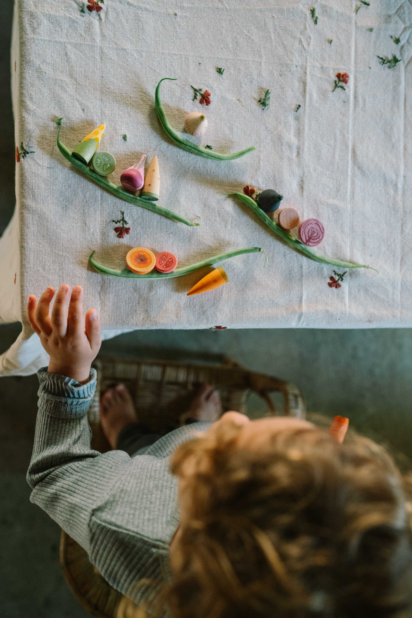 A child arranges the Grapat Play Set - Veggies, with wooden green beans, carrot slices, and colorful pieces on a white floral tablecloth. Their hand and curly hair are visible as they enjoy imaginative play.