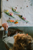 A child arranges the Grapat Play Set - Veggies, with wooden green beans, carrot slices, and colorful pieces on a white floral tablecloth. Their hand and curly hair are visible as they enjoy imaginative play.