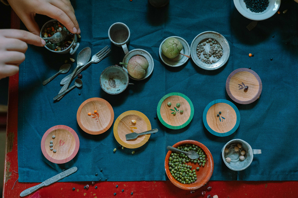 The Grapat Play Set - Dishes features a vibrant arrangement of seeds and spices in small bowls, wooden plates, and trays on a blue cloth. Hands use spoons and cups to sort, with scattered spoons and loose seeds creating a playful scene.