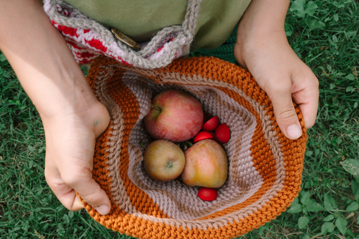 A person stands on green grass, holding a knitted hat filled with freshly picked apples, small red berries, and Grapat Mandala Half Spheres.