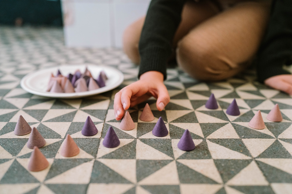 A child arranges small purple and mauve Grapat Mandala Peak toys, made from sustainable wood, into patterns on a geometric floor, with more cone-shaped pieces on a nearby white plate.