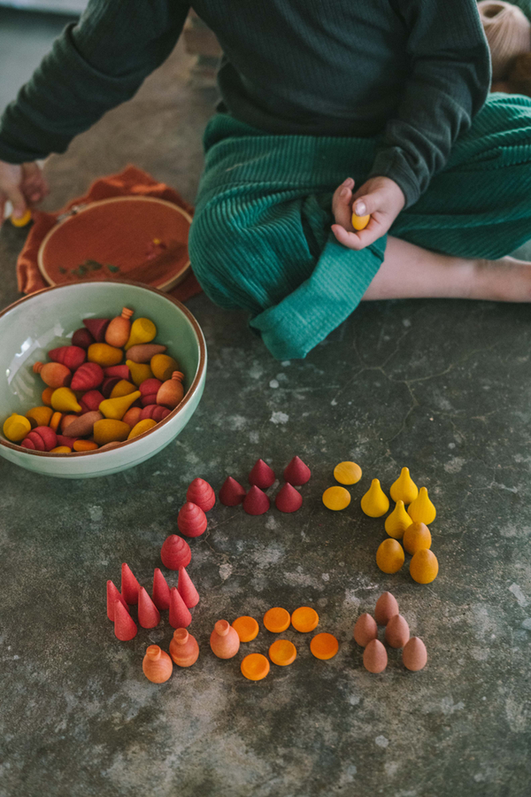 A child in green pants sits on the floor sorting colorful Grapat Mandala Mix - Warm wooden pieces—red, yellow, and orange—into groups on a gray surface, with bowls and a plate placed nearby.