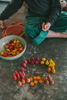 A child in green pants sits on the floor sorting colorful Grapat Mandala Mix - Warm wooden pieces—red, yellow, and orange—into groups on a gray surface, with bowls and a plate placed nearby.