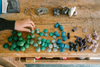 A hand reaches for the Grapat Mandala Mix - Cold, small sustainable wooden toys in cool shades of green, blue, purple, and tan arranged by color on a rustic table, highlighting their enchanting charm.